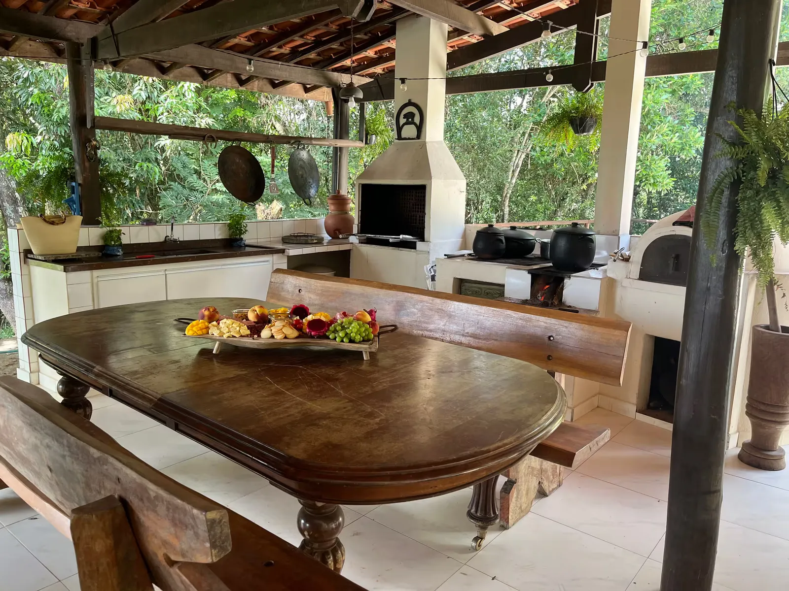 Espaço gourmet coberto do Sítio Manacás com mesa oval de madeira, churrasqueira e forno a lenha