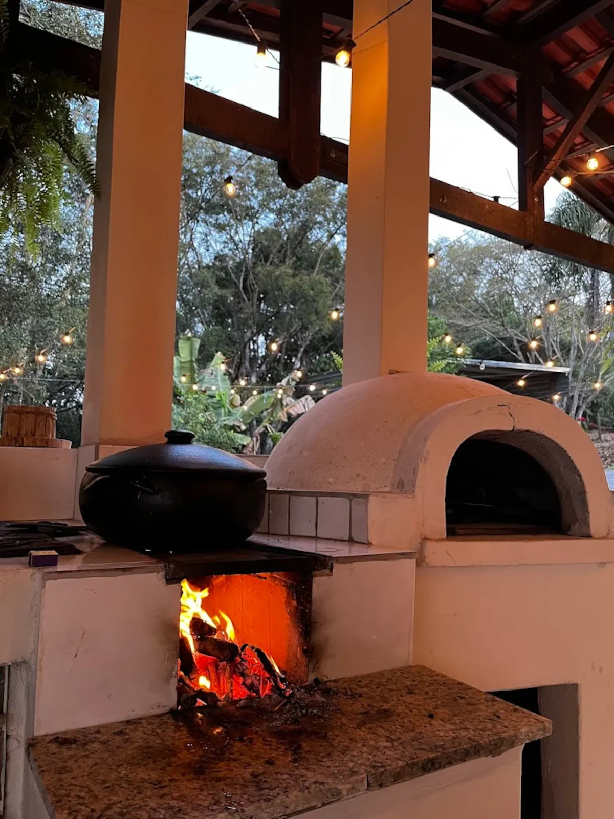Forno a lenha aceso no espaço gourmet do Sítio Manacás com varal de luzes ao anoitecer