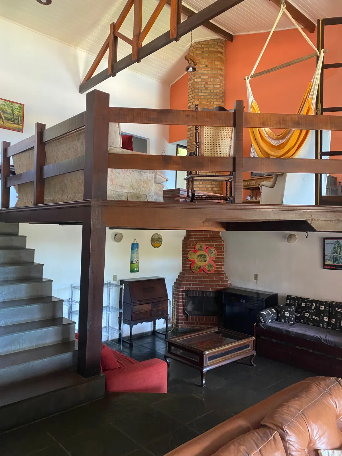 Sala de estar da casa sede do Sítio Manacás com lareira de tijolos, mezanino de madeira e rede suspensa